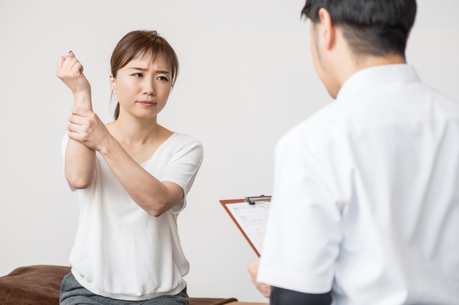 顔をしかめて手首を押さえる女性と、バインダーを持つ男性