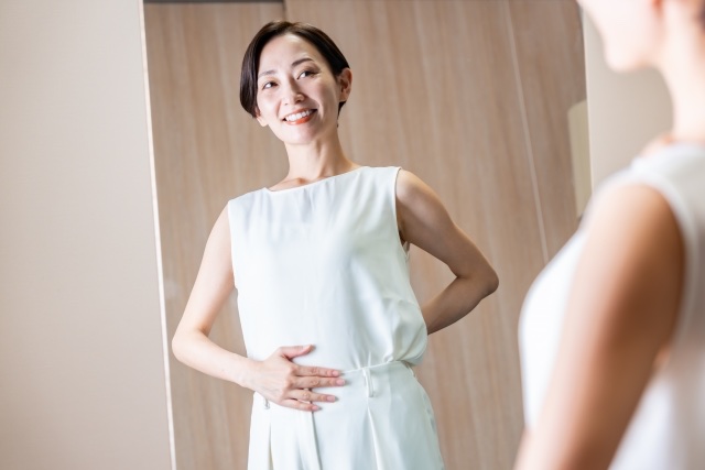 鏡に映る白いノースリーブの女性が腰とおなかに手を当てる