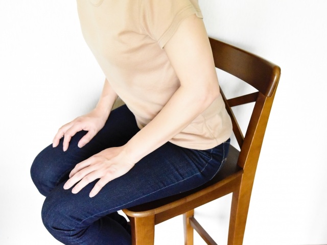 木の椅子の前のほうに浅く腰かけ、両手を膝に置く女性