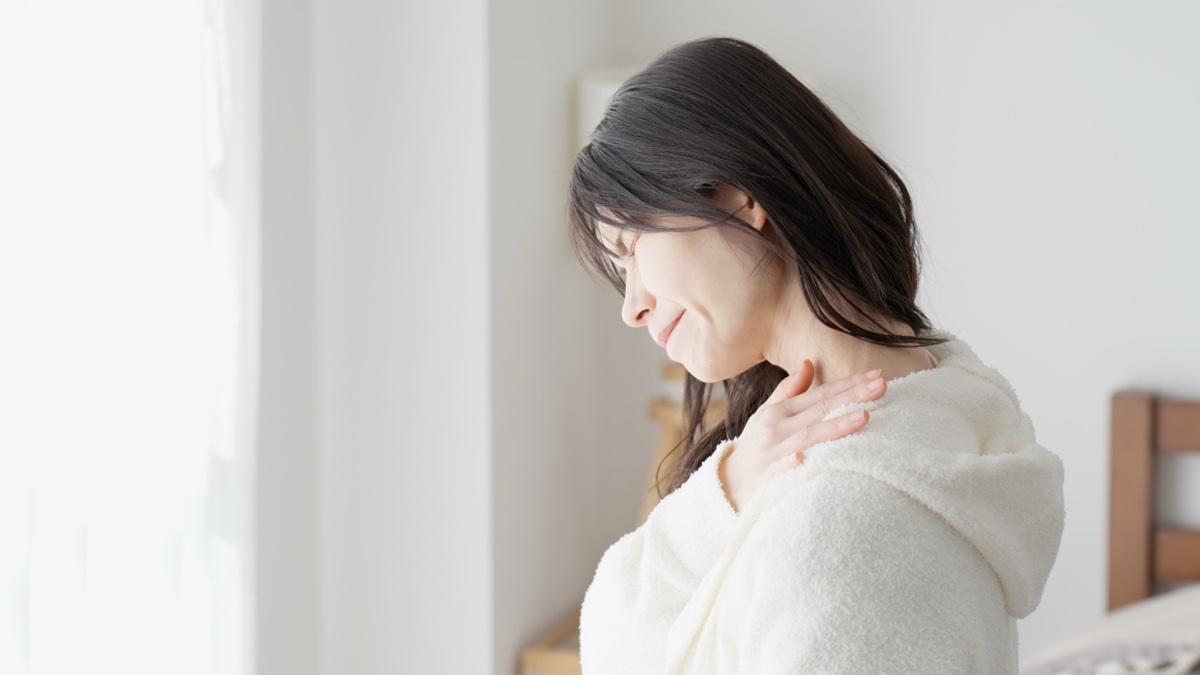 窓辺で白いルームウェアの女性が肩に手を当てる